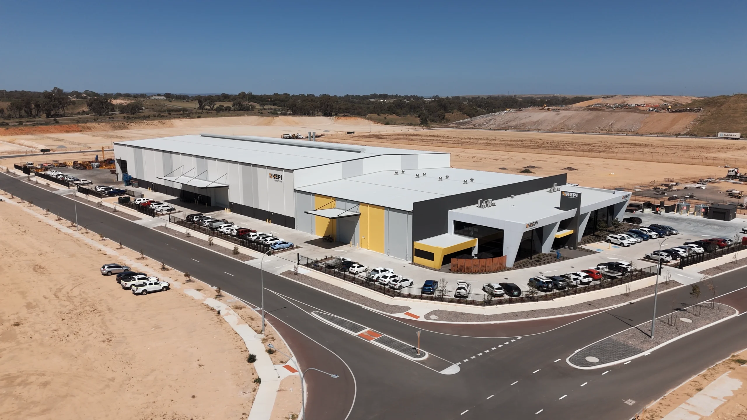 Aerial view of a modern industrial building complex with white and gray façades, yellow accents, and a large parking lot, set in a sandy desert landscape under a clear blue sky.