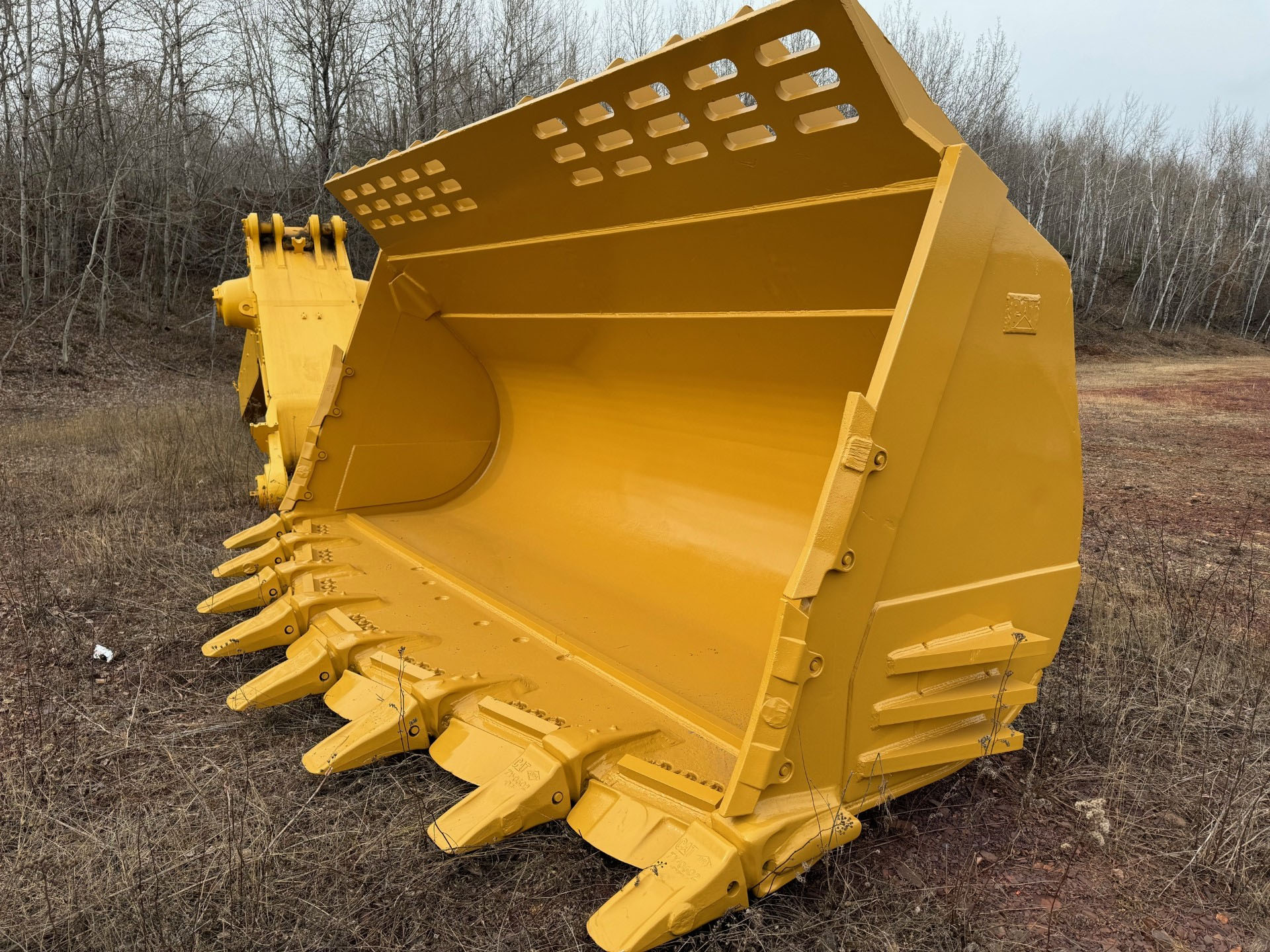 Yellow excavator bucket attachment resting in a dry grassy field at the edge of a bare-wooded area.