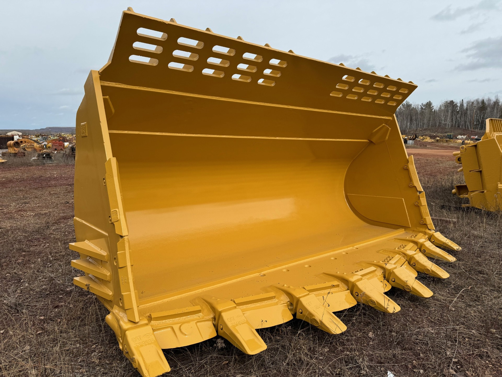 Close-up of a large yellow excavator bucket attachment in a salvage yard with other machinery in the background.