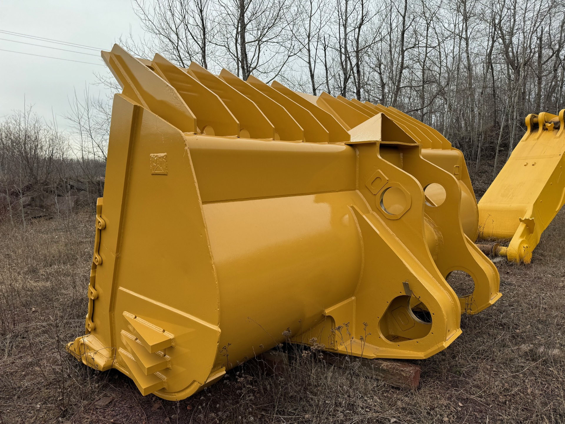 Large yellow excavator bucket attachment resting on dry grass in a bare-wooded area.