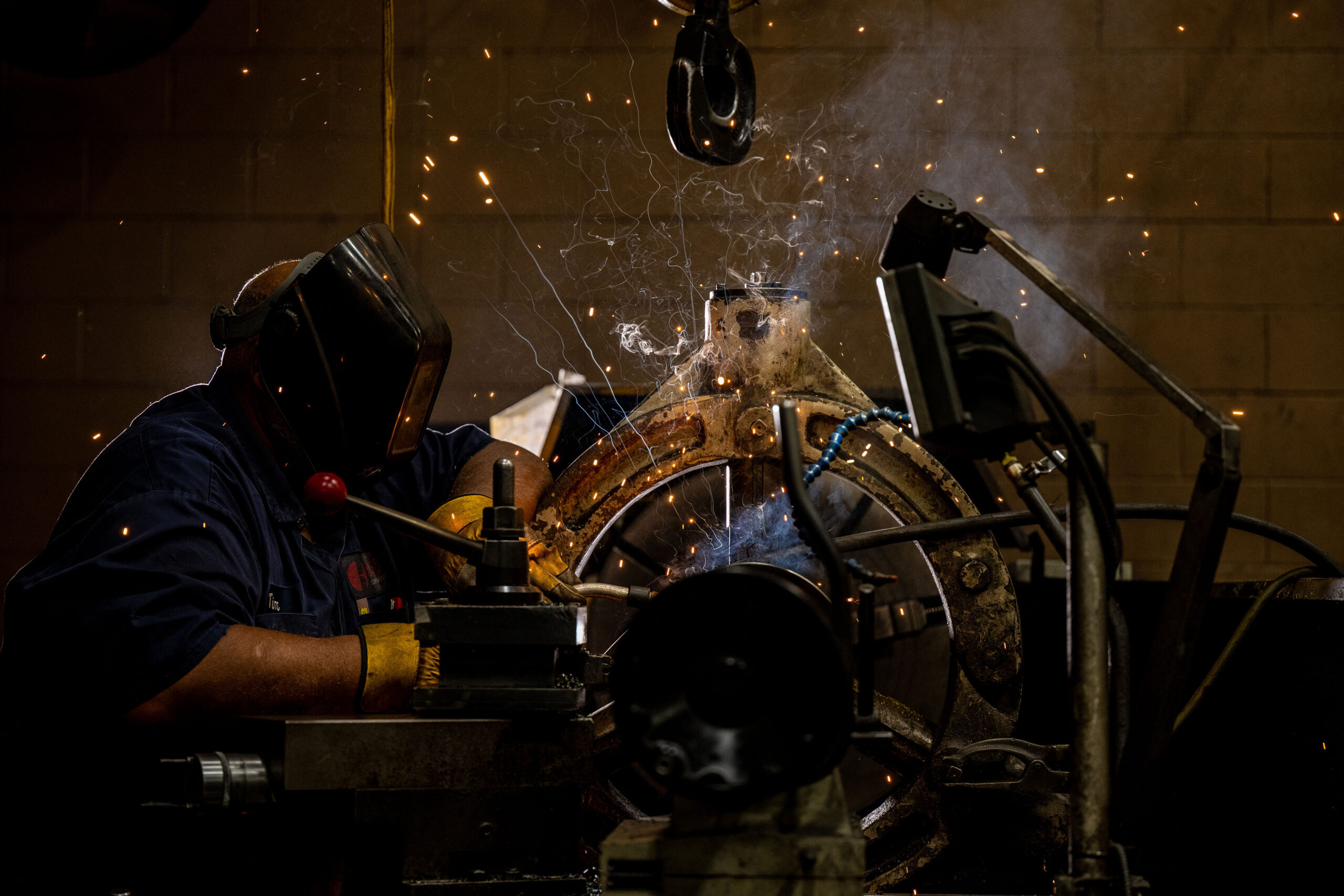 Welder in protective gear working on metal, sparks flying as they weld a large joint in a dim workshop.