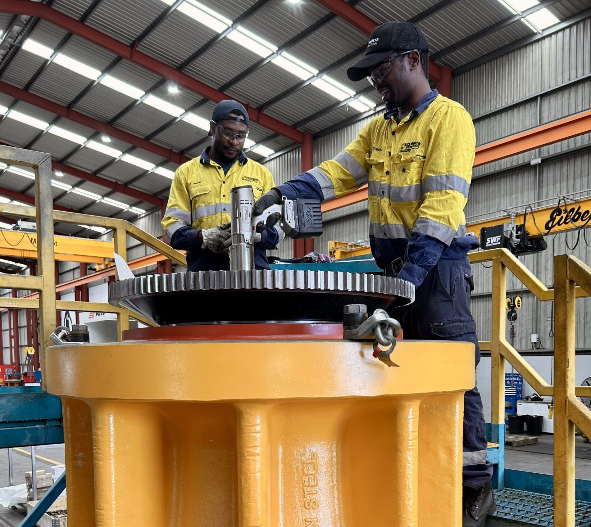 Two workers in yellow safety jackets and hard hats assemble a large yellow gear in an industrial workshop.