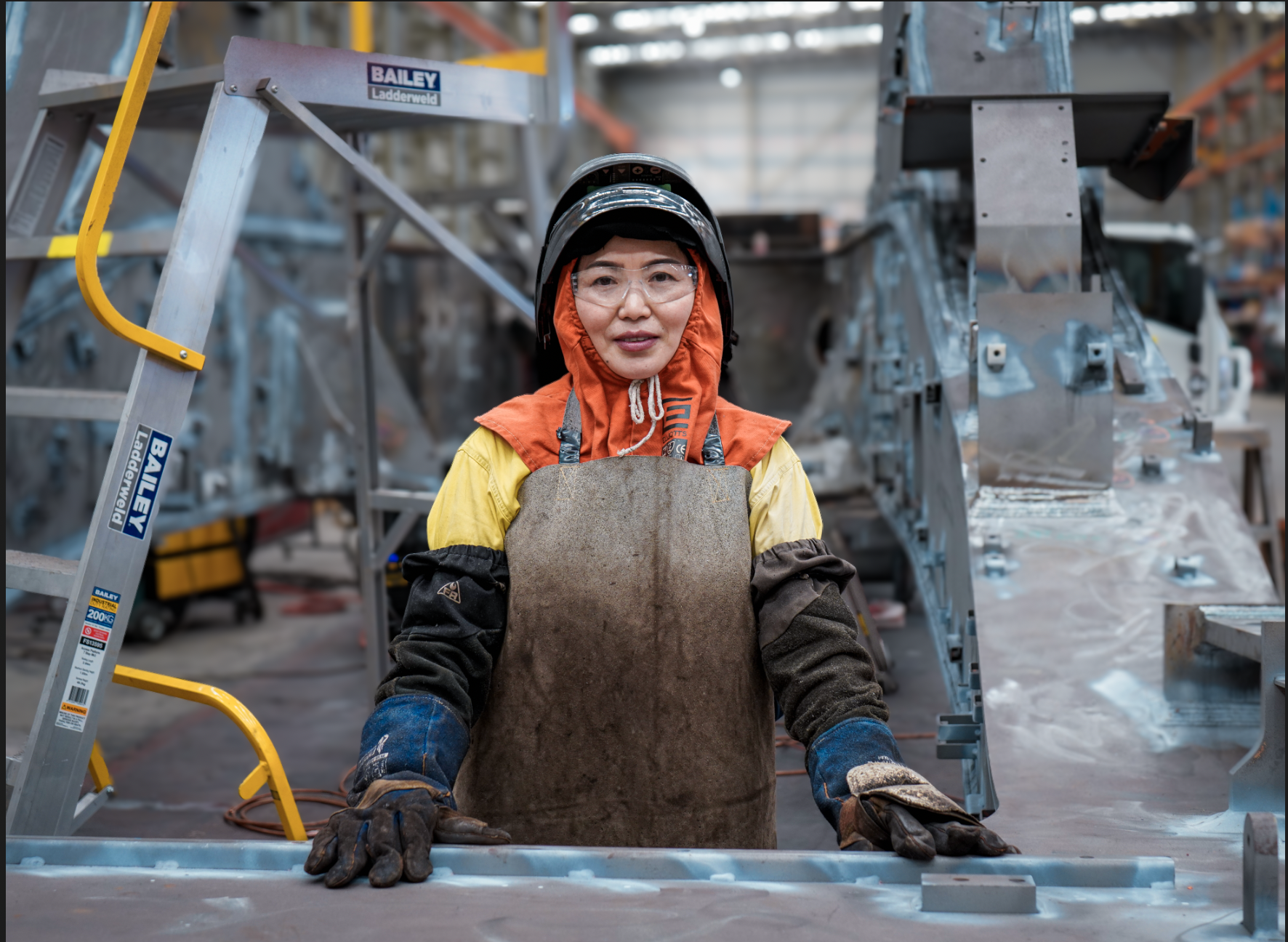 Female factory worker in a helmet and safety goggles stands at a metal workstation in an industrial plant with equipment around.